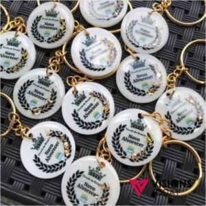White round keychains with gold chains, each showing a laurel wreath and the text 'Novo Alvorecer' with a small colorful emblem in the center (brand badge).